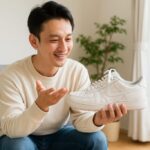 明るい部屋でソファに座る日本人男性が、手にした白いスニーカーを眺めながら微笑んでいる様子。