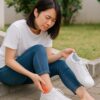 スニーカーを脱いで足首の後ろを押さえ、靴擦れの痛みに顔をしかめる日本人女性が屋外の階段に座っている様子。
