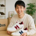 明るい部屋で日本人男性がモーブスのスニーカーを手に持ち、笑顔で座っている様子。テーブルの上にはスニーカーの箱も置かれており、清潔感のある雰囲気が伝わる。