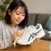 日本人女性が室内でアシックスの白いスニーカーを丁寧に手入れしている様子。明るく清潔感のある部屋で、スプレーボトルと布を使いながら加水分解を防ぐケアを行っている。