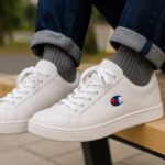 白のチャンピオンスニーカーを日本人がデニムとグレーの靴下に合わせて履き、屋外のベンチで足元を写した横長構図の写真。