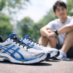 公園の明るい日差しの中、ASICS GEL-CUMULUS16のスニーカーが地面に置かれ、背景には座ってくつろぐ日本人男性がやわらかくボケて写っているシーン